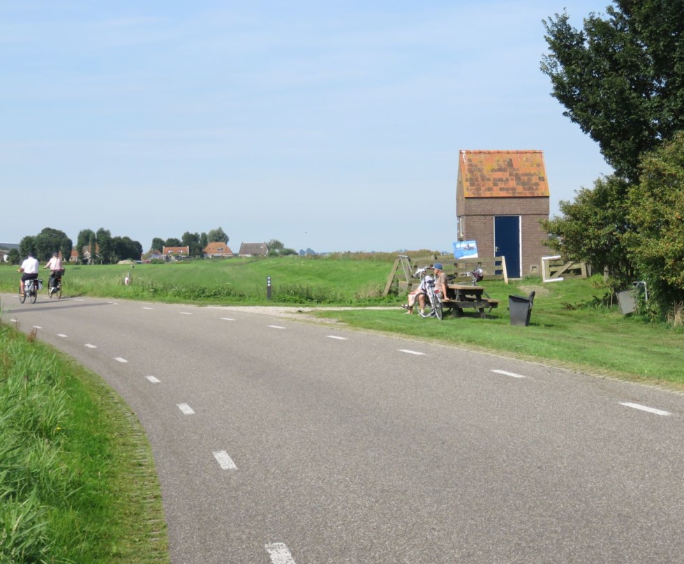 Foto van een gemaal met een weg ervoor met fietsers. In de verte zijn nog huizen te zien